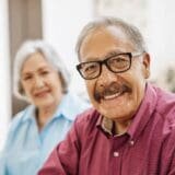 An older male adult smiling at the camera at his house with his wife blurry in the background